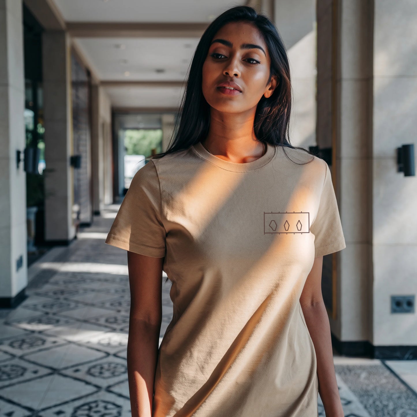 Woman standing inside a modern art museum wearing a Beige Supima cotton T-shirt by Lilak, featuring a minimal geometric design on the left chest