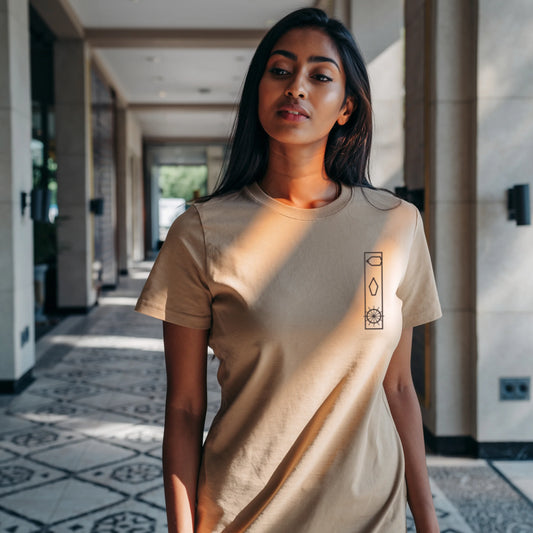 Woman walking down a sunlit city street wearing a light beige Lilak T-shirt with a minimal vertical graphic on the left chest, paired with blue 