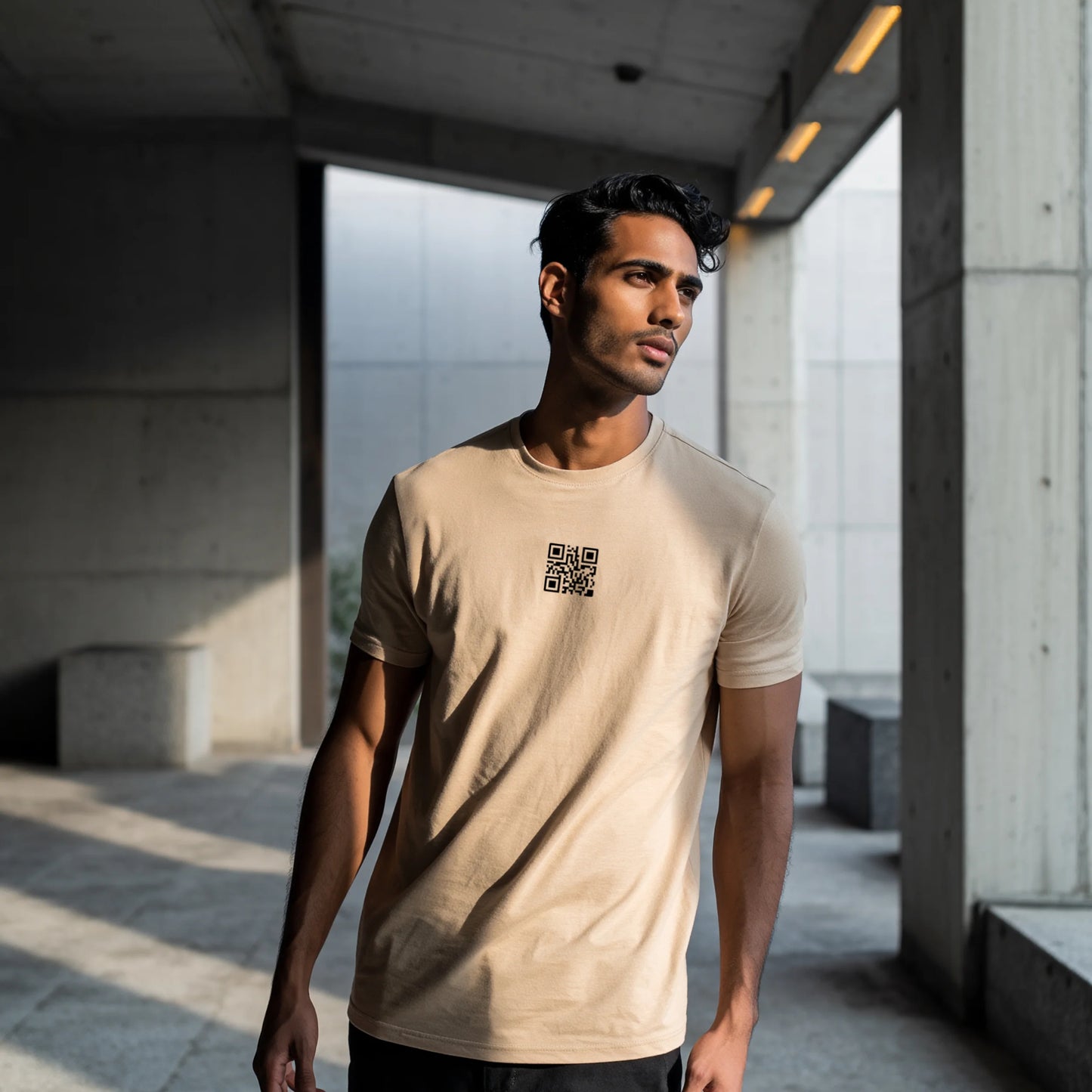Man wearing a Beige T-shirt with a black QR code printed at the center of the chest, standing indoors in soft natural light with hands in pockets