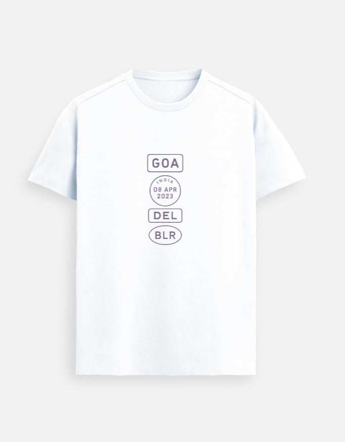 Indian male model wearing a white Supima cotton T-shirt by Lilak, featuring a minimalistic violet travel-stamp design reading GOA, INDIA 08 APR 2023, DEL, and BLR. Model stands against a textured grey studio background