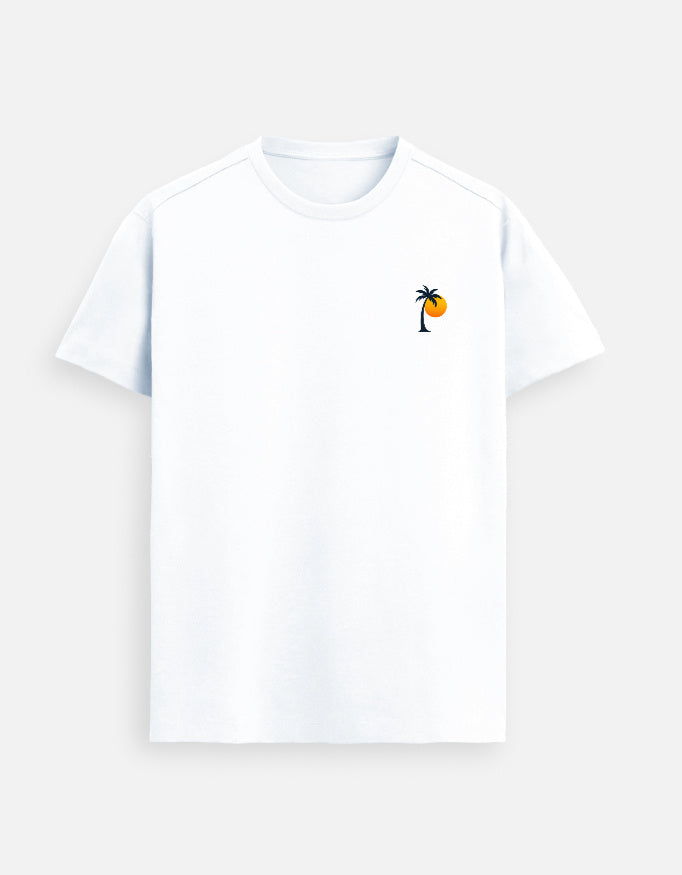 Woman sitting at a wooden table holding a cup, wearing a White Lilak T-shirt with a small palm tree and sun graphic on the left chest, in warm sunset lighting against a minimal beige background