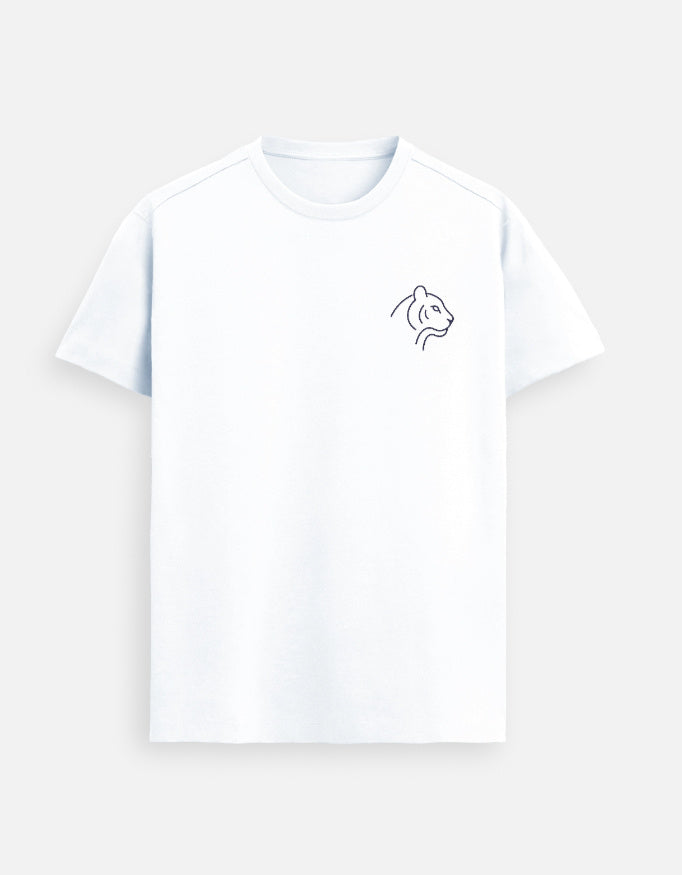 Woman wearing a White Lilak T-shirt with a small minimal Tiger illustration on the left chest, seated at an outdoor café table in a lively, tree-lined street setting with warm evening lights