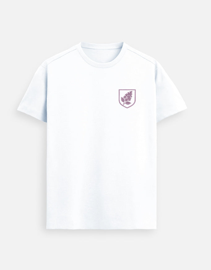 Female model wearing a White Supima cotton T-shirt by Lilak featuring a small maroon botanical crest on the left chest, standing in a softly lit studio with large window shadows