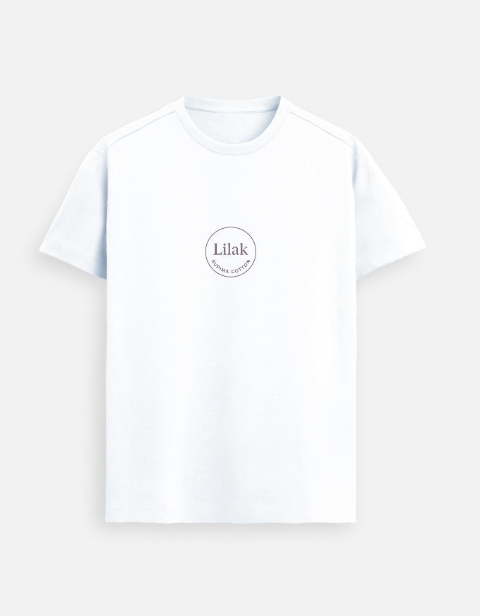 Smiling man wearing a White Lilak Supima cotton T-shirt with a circular logo on the chest, standing in a softly lit modern room with wooden furniture and indoor plants