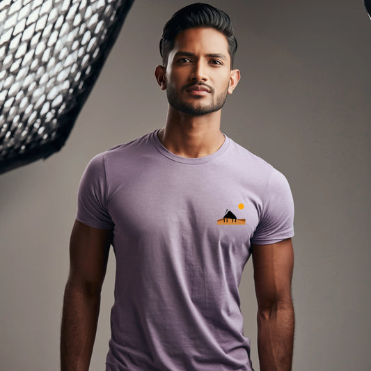 Male model standing in a softly lit studio wearing a Lavender Supima cotton T-shirt by Lilak, featuring a minimal sunset hut graphic on the left chest