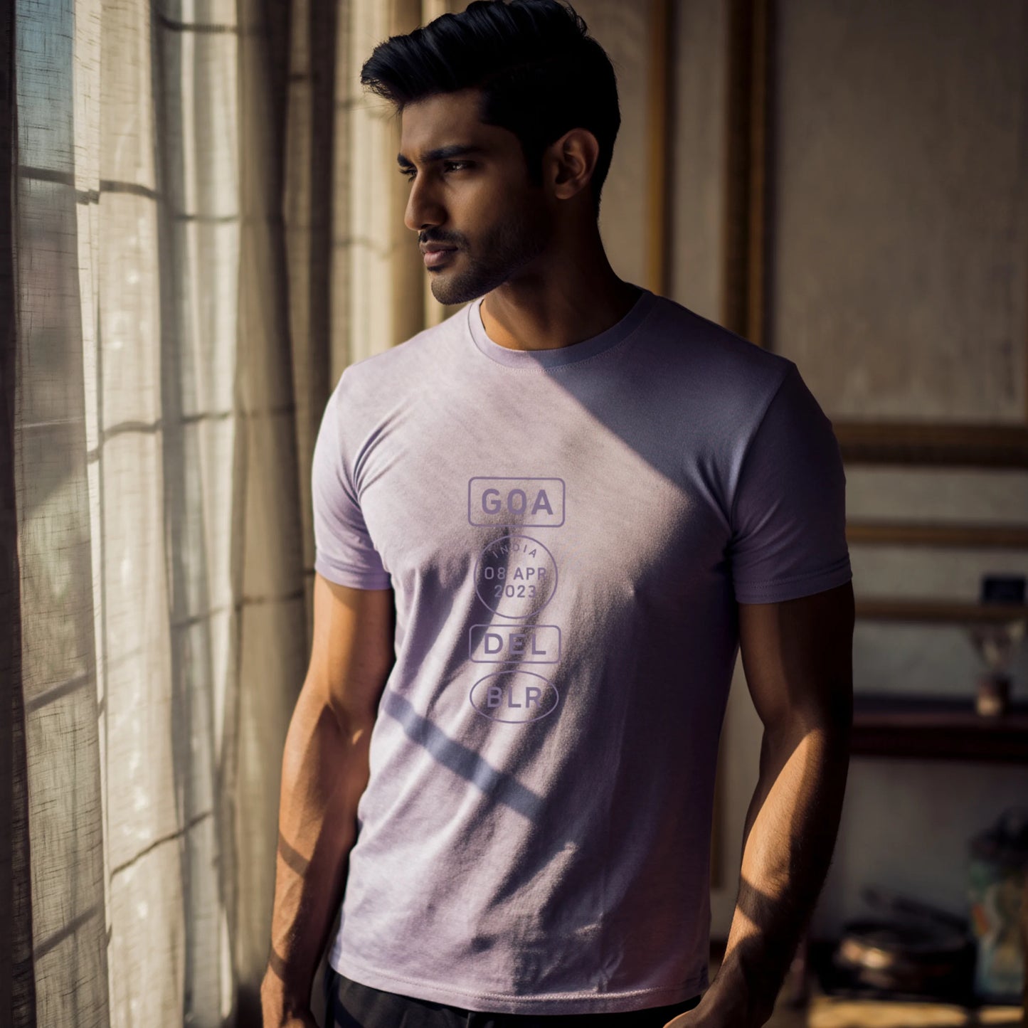 Indian male model wearing a Lavender Supima cotton T-shirt by Lilak, featuring a minimalistic violet travel-stamp design reading GOA, INDIA 08 APR 2023, DEL, and BLR. Model stands against a textured grey studio background