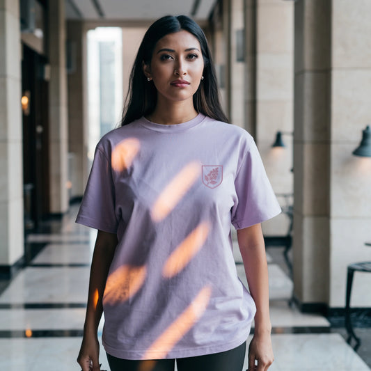 Female model wearing a Lavender Supima cotton T-shirt by Lilak featuring a small maroon botanical crest on the left chest, standing in a softly lit studio with large window shadows