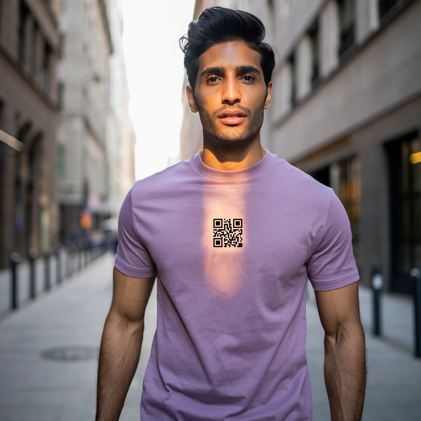 Man wearing a Lavender T-shirt with a black QR code printed at the center of the chest, standing indoors in soft natural light with hands in pockets