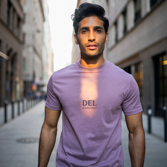 Male model standing indoors wearing a Lavender Lilak Supima cotton T-shirt with ‘DEL’ and geographic coordinates printed on the chest, photographed in soft natural light with a plant and wooden furniture in the background