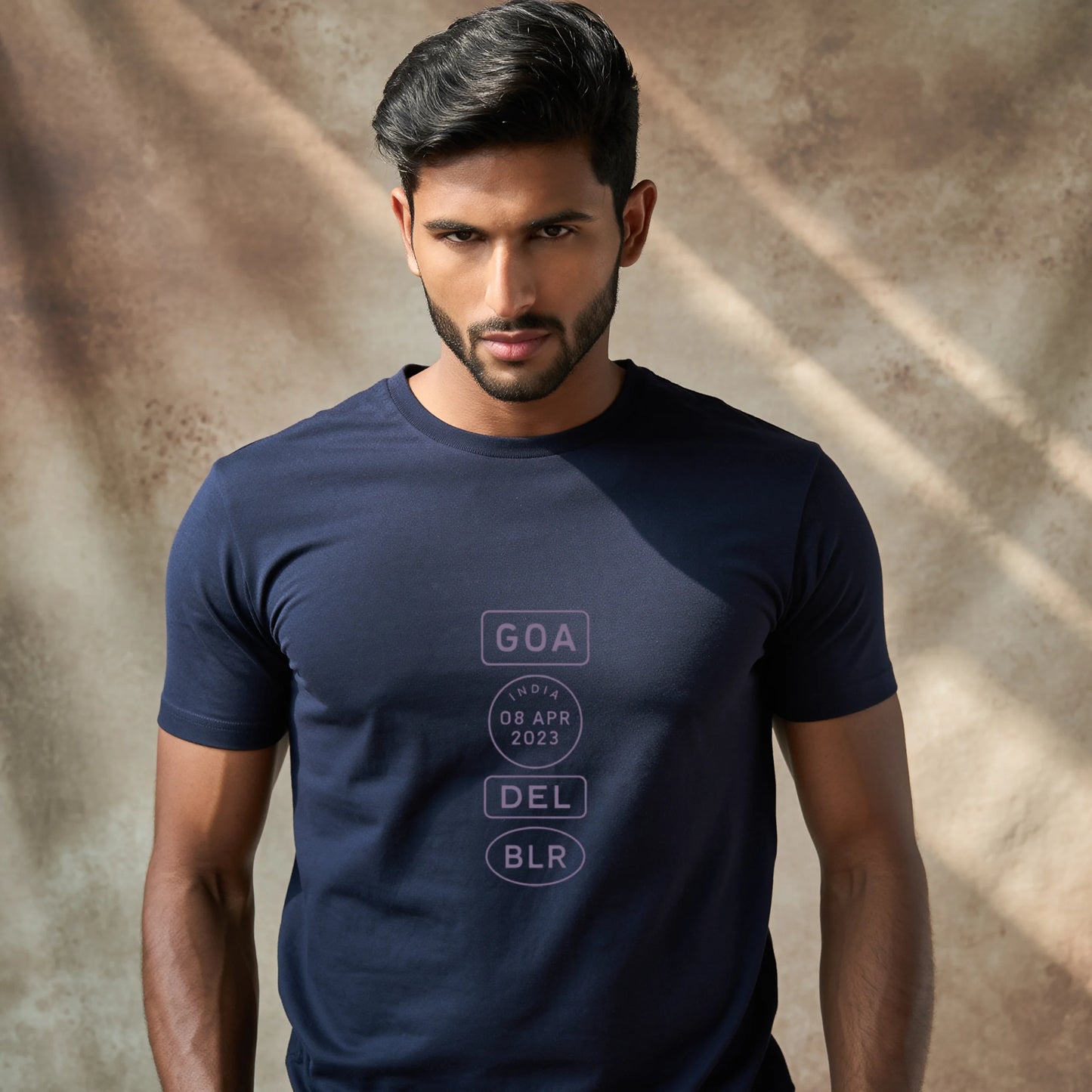 Indian male model wearing a Navy Blue Supima cotton T-shirt by Lilak, featuring a minimalistic violet travel-stamp design reading GOA, INDIA 08 APR 2023, DEL, and BLR. Model stands against a textured grey studio background