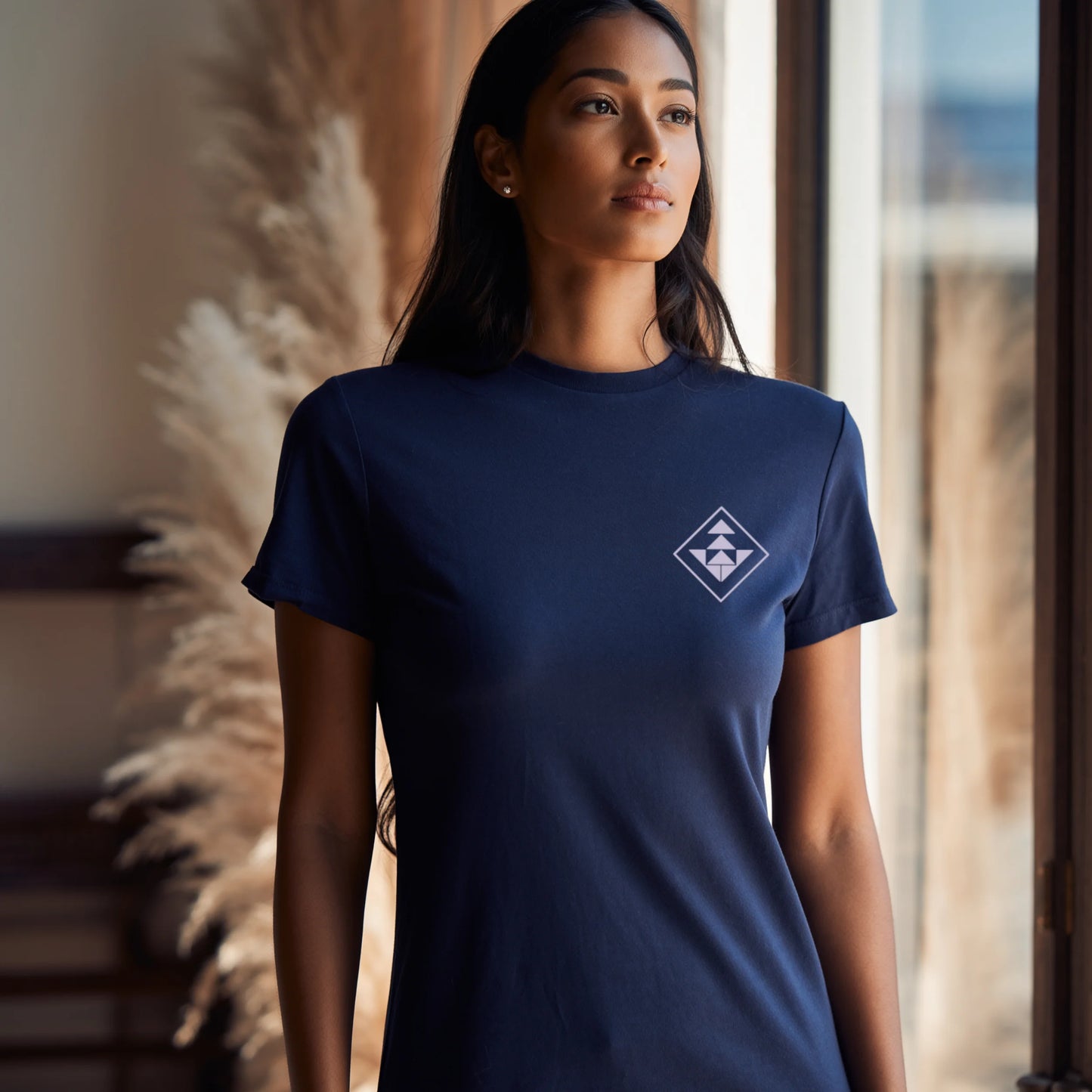 Woman standing in a stadium lounge wearing a Navy Blue Lilak T-shirt with a purple geometric emblem on the left chest, with sunlight shining over the empty cricket ground in the background