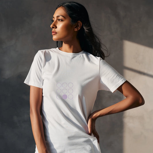 Indian female model wearing a White Supima cotton T-shirt by Lilak, featuring a minimal lavender geometric circle design on the center chest. She stands in a softly lit studio with large window shadows in the background