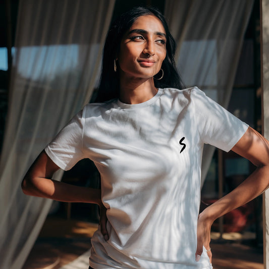 Woman wearing a White Lilak T-shirt featuring a small black minimalist ‘S’ symbol on the left chest, standing indoors in soft natural light with a calm expression