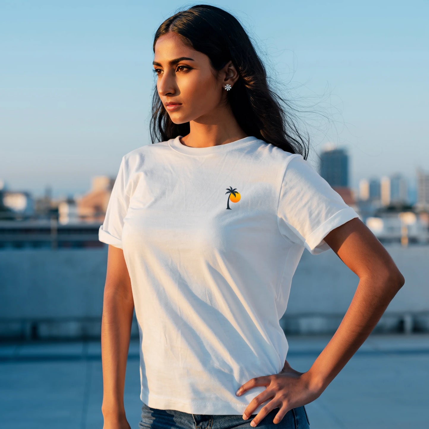Woman sitting at a wooden table holding a cup, wearing a White Lilak T-shirt with a small palm tree and sun graphic on the left chest, in warm sunset lighting against a minimal beige background