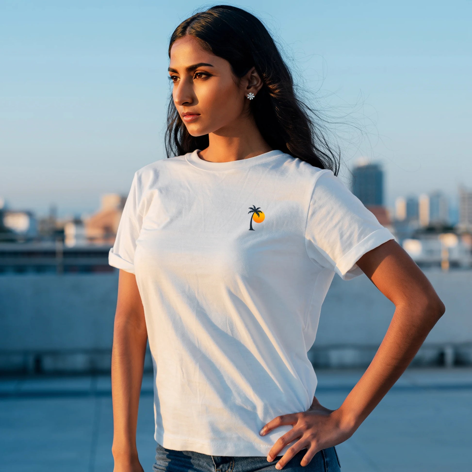 Woman sitting at a wooden table holding a cup, wearing a White Lilak T-shirt with a small palm tree and sun graphic on the left chest, in warm sunset lighting against a minimal beige background